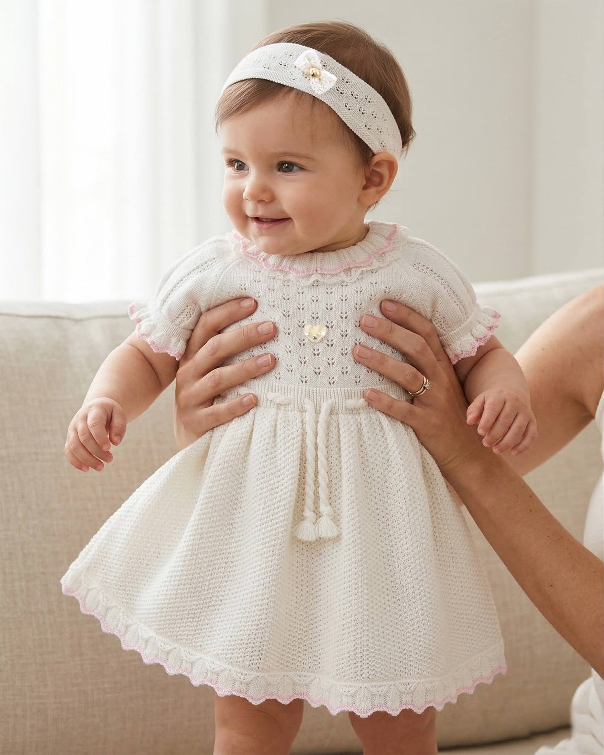 Baby in a white lace dress held by an adult, sitting on a couch.
