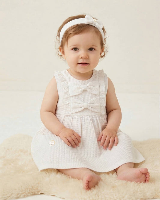 Baby in a white dress with a bow sitting on a beige surface