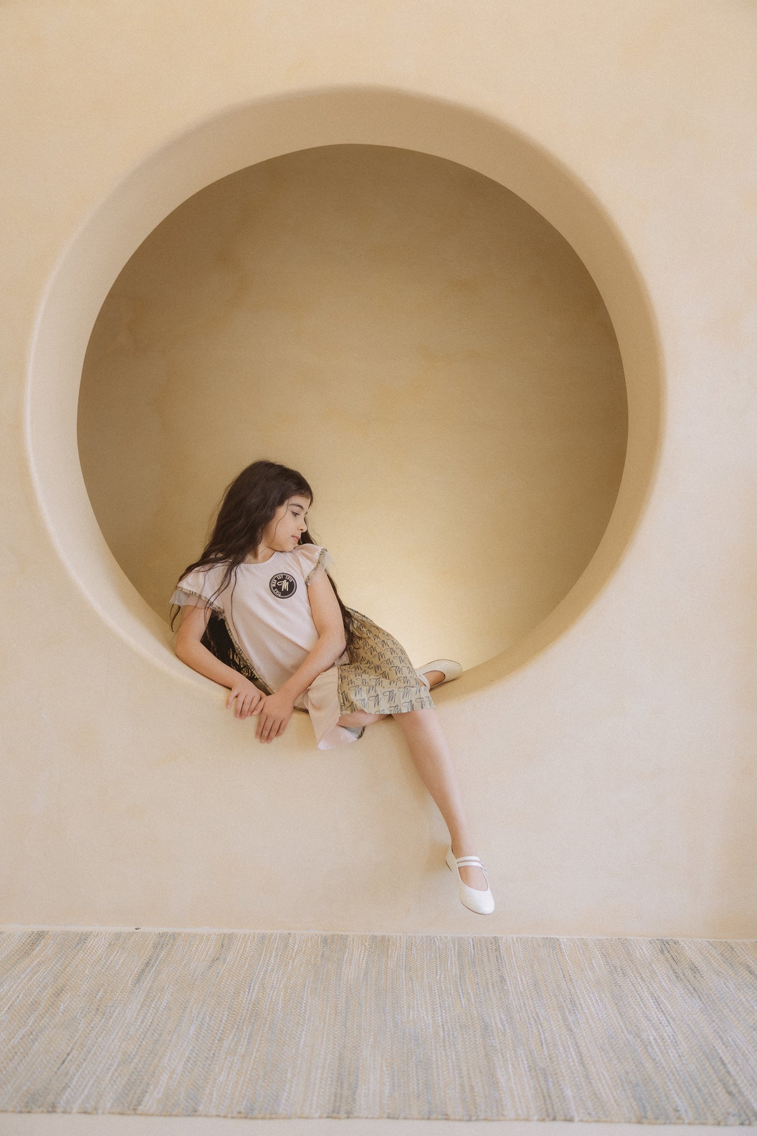 A young girl sitting inside a round niche, wearing a pink dress with contrast side panels and a unique script print, short ruffle sleeves, and a emblem on the chest.