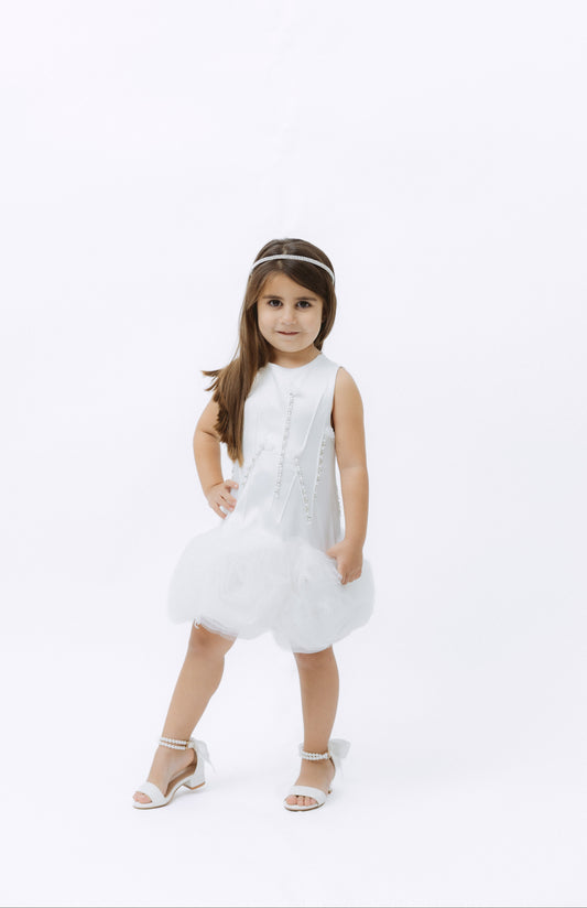 A young girl with long brown hair stands on a seamless white studio background wearing a sleeveless white dress with a tulle tutu skirt, a white headband, and white heeled sandals.
