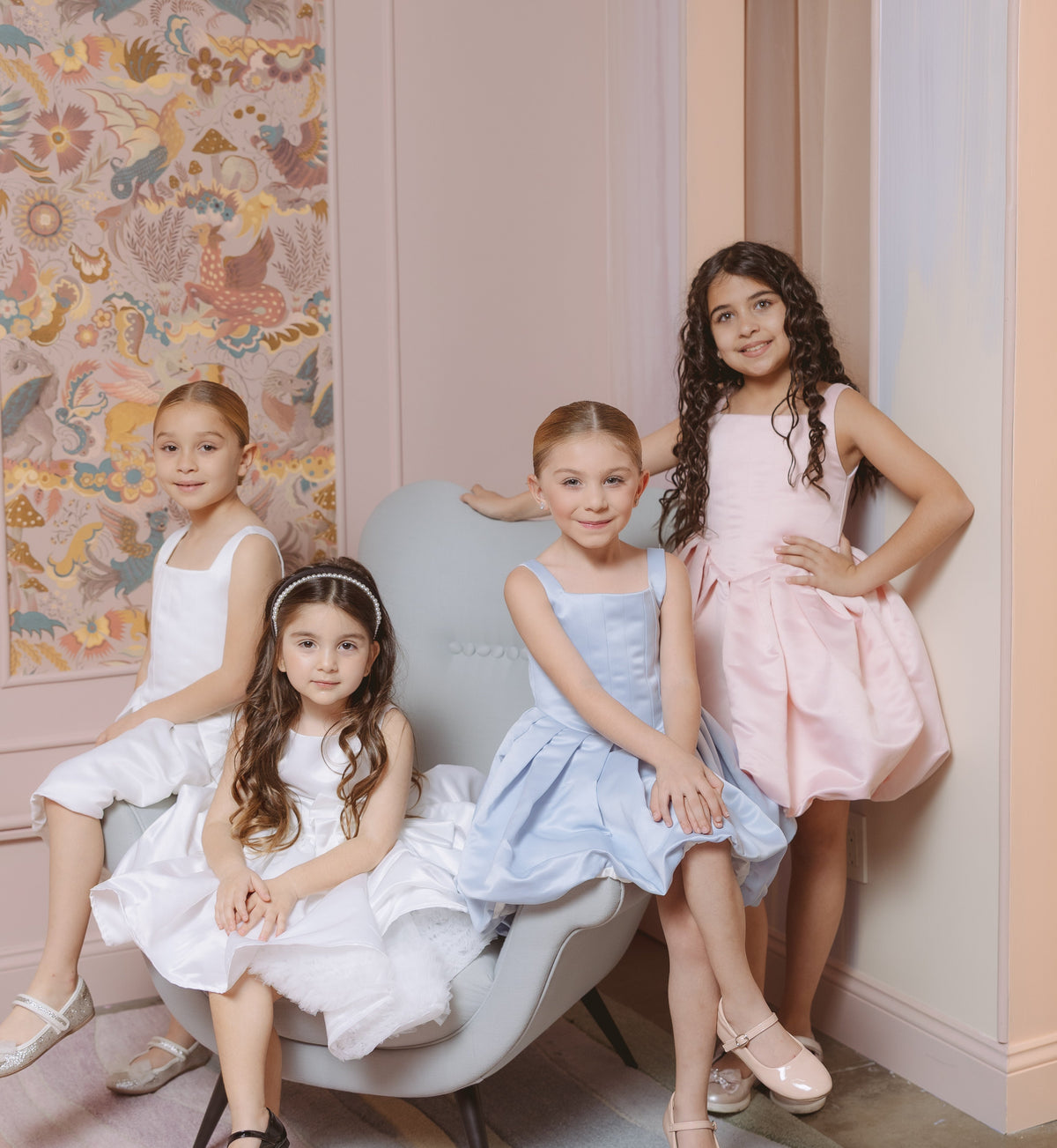 Four young girls in dresses posing in a room with patterned wallpaper.