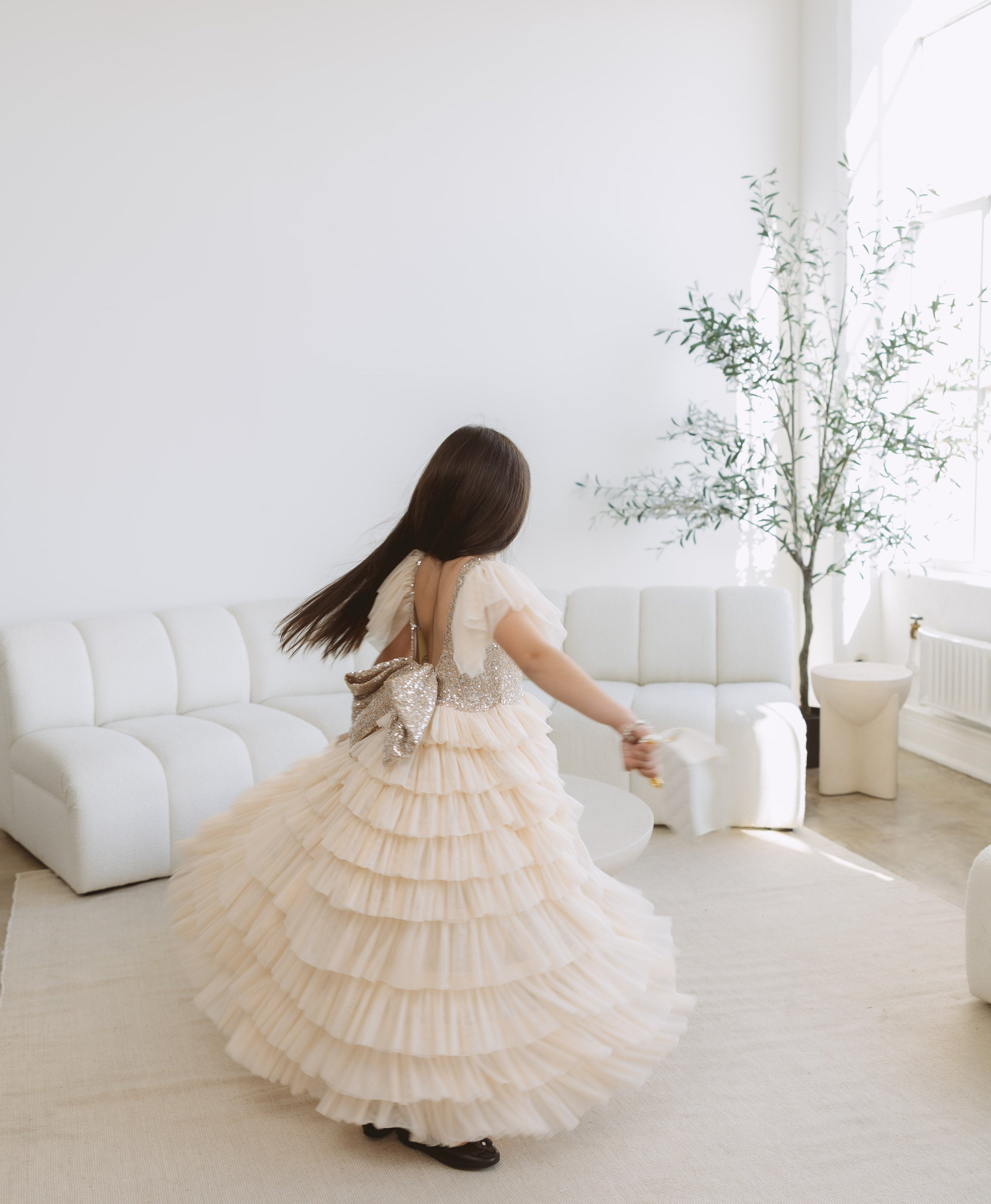 A young girl with long dark hair is seen from behind, wearing a voluminous cream-colored ruffled ball gown and twirling in a bright, modern white living room.